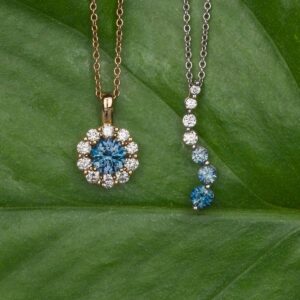 Two "Necklaces" with blue and white gemstones rest on a green leaf: one features a round halo pendant, while the other displays a vertical line of alternating gems.