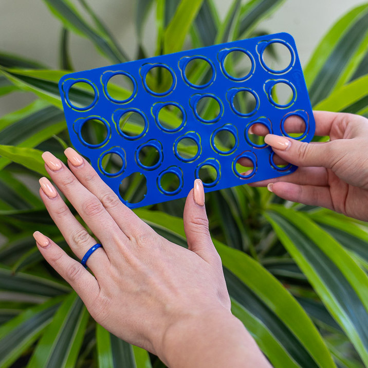 A person uses the blue Ring-Sizer tool to try on a blue ring, possibly checking sizes for engagement rings, with green leaves in the background.