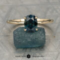 The Royea, Alexander gold solitaire ring with an oval-cut dark blue gemstone rests atop a rough blue stone, "Earth's Treasury" logo below. This Hidden Accent Solitaire radiates subtle elegance and timeless charm.