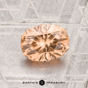 The 1.44-Carat Grossular Garnet, cut in a faceted oval shape and peach-colored, is shown on a textured metallic surface with "EARTH'S TREASURY" at the bottom.