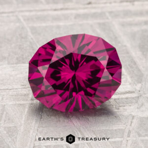 A 2.75-carat Umbalite Garnet, faceted in an oval cut and displaying a deep magenta hue, rests on a textured light gray surface with "EARTH'S TREASURY" shown at the bottom.