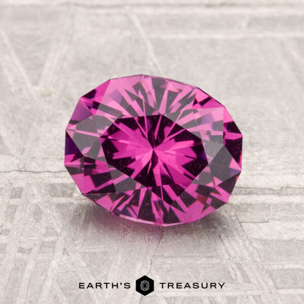 1.90-Carat Mahenge Garnet A vivid, faceted oval 1.90-carat Mahenge Garnet gemstone is showcased on a textured, light surface with "EARTH'S TREASURY" inscribed below.