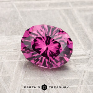 A 1.63-carat Mahenge Garnet, faceted in an oval cut, is displayed on a textured silver-gray surface with the text "EARTH'S TREASURY" below.
