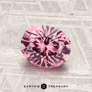 A 1.59-carat faceted oval Mahenge garnet is displayed on a textured light gray surface, with the text "EARTH'S TREASURY" at the bottom.