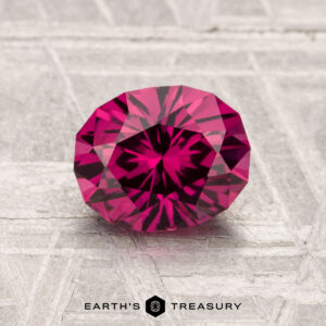 A 1.57-carat Mahenge Garnet, cut in a round brilliant style, is showcased on a textured gray surface with "EARTH'S TREASURY" at the bottom.