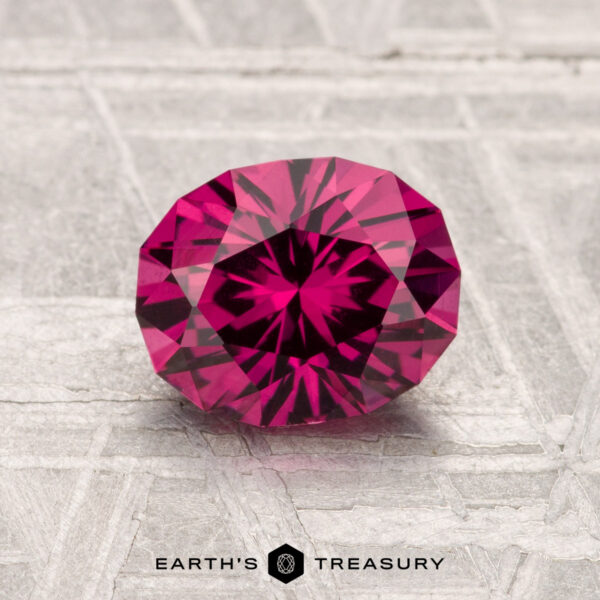 1.57-Carat Mahenge Garnet A 1.57-carat Mahenge Garnet, cut in a round brilliant style, is showcased on a textured gray surface with "EARTH'S TREASURY" at the bottom.