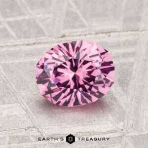 A 1.38-carat Mahenge Garnet, round brilliant-cut, is displayed on a textured light gray surface with "EARTH'S TREASURY" at the bottom.