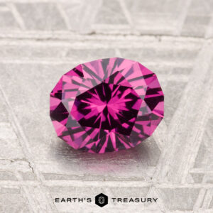 A 1.38-carat Mahenge Garnet, round and faceted, sits on a textured silver background with "EARTH'S TREASURY" displayed below.