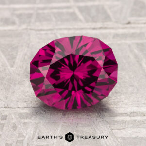 A 2.33-carat oval Umbalite garnet with a deep pink hue rests on a textured gray surface, "EARTH'S TREASURY" displayed at the bottom.