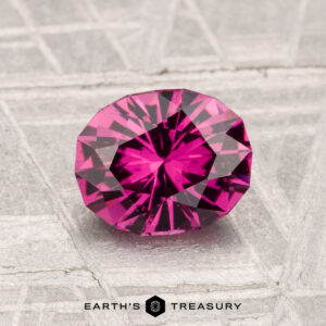 A 1.35-carat Mahenge Garnet, cut in an oval and displaying a vivid pink hue, rests on a textured gray surface with the text "EARTH'S TREASURY" below.
