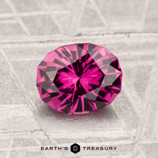 1.35-Carat Mahenge Garnet A 1.35-carat Mahenge Garnet, cut in an oval and displaying a vivid pink hue, rests on a textured gray surface with the text "EARTH'S TREASURY" below.