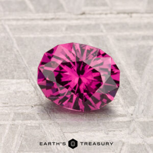 A 1.33-carat Mahenge Garnet, cut in a faceted oval shape, is showcased on a textured gray surface with the "Earth's Treasury" logo at the bottom.