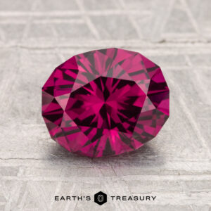 2.28-Carat Umbalite Garnet, oval-cut and deep pink, shown on a textured gray surface with the "Earth's Treasury" logo and text at the bottom.