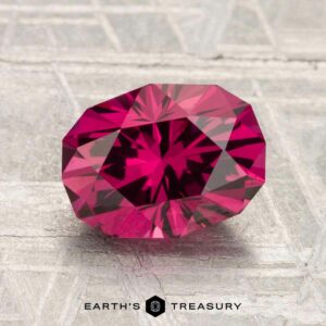 A faceted 2.50-Carat Mahenge Garnet, oval-cut and raspberry pink, is showcased on a textured light surface with "Earth's Treasury" below, highlighting its brilliance.
