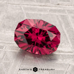 A faceted 2.37-carat Mahenge Garnet with a deep red hue rests on a textured silver-gray surface, with "Earth's Treasury" at the bottom.