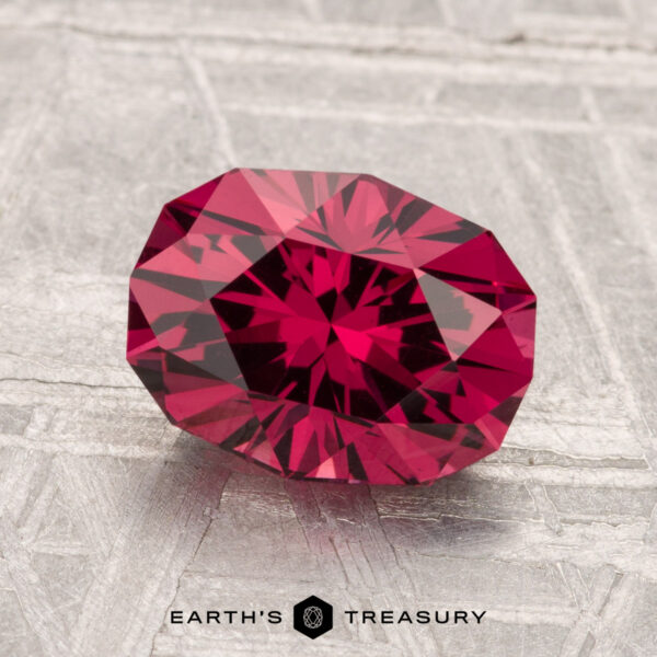 2.37-Carat Mahenge Garnet A faceted 2.37-carat Mahenge Garnet with a deep red hue rests on a textured silver-gray surface, with "Earth's Treasury" at the bottom.