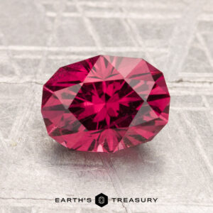 A 2.24-carat Mahenge garnet, faceted in an oval shape, rests on a textured surface with "EARTH'S TREASURY" displayed at the bottom.