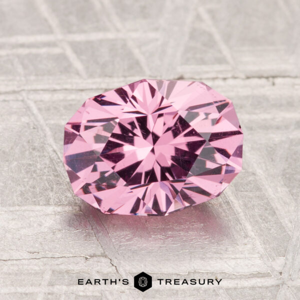 1.88-Carat Mahenge Garnet A 1.88-carat faceted oval Mahenge Garnet in light pink rests on a textured metallic surface, with "EARTH'S TREASURY" displayed at the bottom.