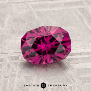 A 1.58-carat Mahenge Garnet, with a faceted deep pink hue, sits atop a textured gray surface displaying "EARTH'S TREASURY" at the bottom.