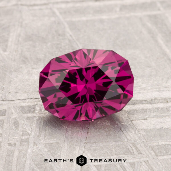 1.58-Carat Mahenge Garnet A 1.58-carat Mahenge Garnet, with a faceted deep pink hue, sits atop a textured gray surface displaying "EARTH'S TREASURY" at the bottom.