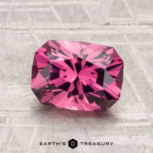 A faceted, rectangular deep pink 2.27-carat Mahenge garnet rests on a textured gray surface; "EARTH'S TREASURY" is written at the bottom.