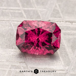 A 2.07-carat rectangular faceted Mahenge Garnet is shown on a textured gray surface with the "Earth's Treasury" logo and text beneath it.