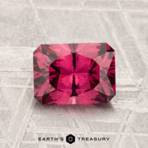 A 1.79-carat Mahenge garnet, cut in a rectangular faceted shape, is shown on a textured light gray surface with the "Earth’s Treasury" text and logo at the bottom.