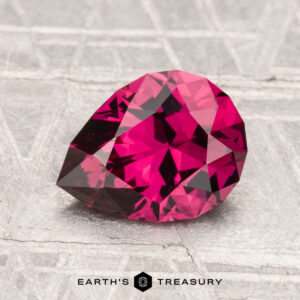 A 1.79-carat, pear-shaped Mahenge Garnet with deep pink facets is displayed on a textured metallic surface, with "Earth's Treasury" below.