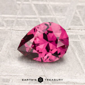 A faceted pear-shaped 1.56-carat Mahenge Garnet gemstone rests on a textured, light-colored surface with the words "EARTH'S TREASURY" below.