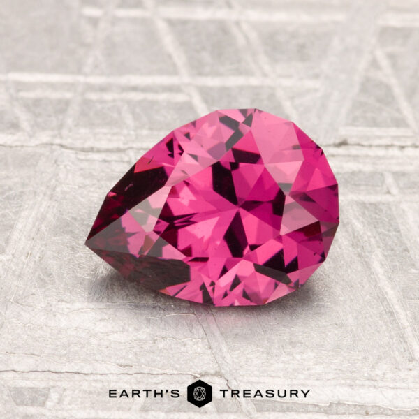 1.56-Carat Mahenge Garnet A faceted pear-shaped 1.56-carat Mahenge Garnet gemstone rests on a textured, light-colored surface with the words "EARTH'S TREASURY" below.