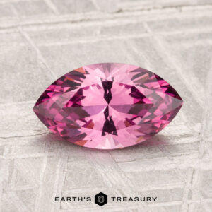A 1.62-carat Mahenge garnet, marquise-cut and pink, rests on a textured light gray surface with “Earth’s Treasury” at the bottom.
