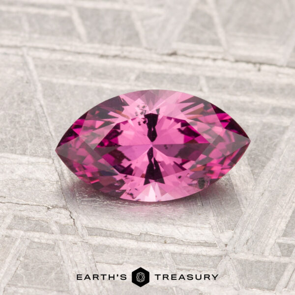 1.17-Carat Mahenge Garnet A 1.17-carat marquise-cut Mahenge Garnet with a vivid pink hue rests on a textured metallic surface, with the text "EARTH'S TREASURY" displayed at the bottom.