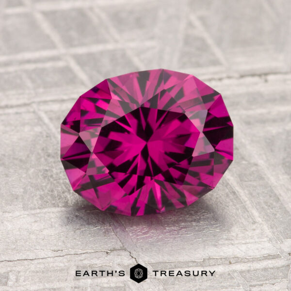 2.24-Carat Umbalite Garnet A 2.24-carat Umbalite Garnet with a deep pink hue is faceted in an oval shape and displayed on a textured gray surface, with "EARTH'S TREASURY" beneath it.