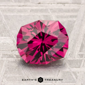 A round, brilliantly faceted 2.49-carat Mahenge garnet gemstone is displayed on a textured surface, with "EARTH'S TREASURY" at the bottom.