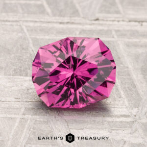 A 1.82-carat Mahenge Garnet, faceted and hot pink, is shown on a textured gray surface with "Earth's Treasury" visible at the bottom.