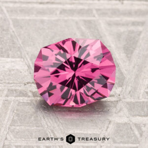 A 1.64-carat Mahenge Garnet, faceted and round with a vibrant pink hue, is displayed on a textured gray surface featuring the "Earth's Treasury" logo at the bottom.