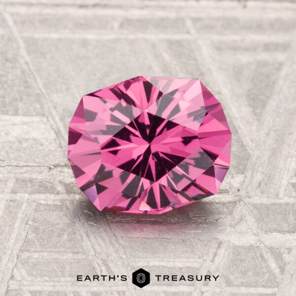 1.64-Carat Mahenge Garnet A 1.64-carat Mahenge Garnet, faceted and round with a vibrant pink hue, is displayed on a textured gray surface featuring the "Earth's Treasury" logo at the bottom.