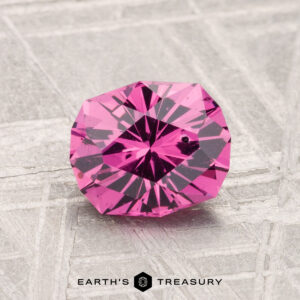 A 1.40-carat, round faceted Mahenge Garnet with a vivid pink hue is shown on a light-textured surface, featuring the "EARTH'S TREASURY" text at the bottom.