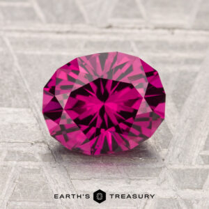 A vivid, round brilliant-cut 2.01-carat Umbalite Garnet displayed on a textured light gray surface with "Earth's Treasury" branding below.
