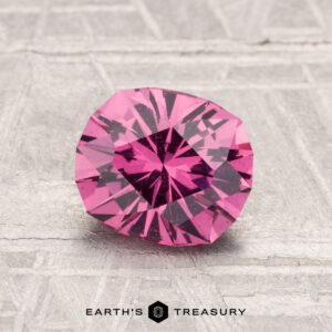 A 1.42-carat Mahenge Garnet, round and faceted with a vivid pink hue, is showcased on a textured gray surface, with "EARTH'S TREASURY" displayed at the bottom.
