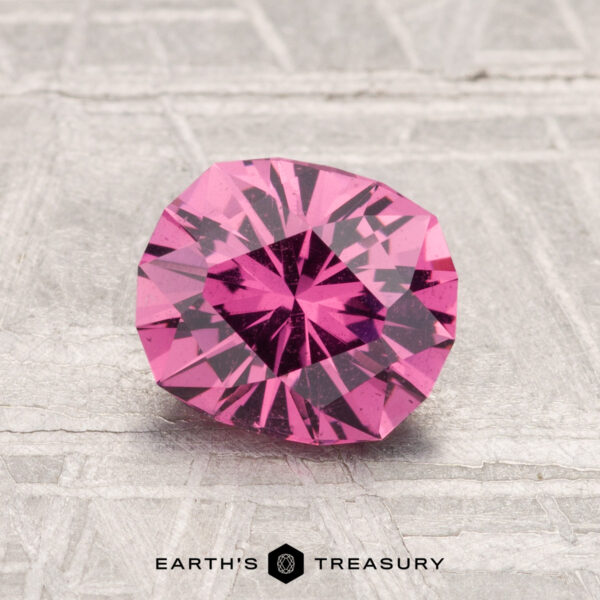 1.42-Carat Mahenge Garnet A 1.42-carat Mahenge Garnet, round and faceted with a vivid pink hue, is showcased on a textured gray surface, with "EARTH'S TREASURY" displayed at the bottom.
