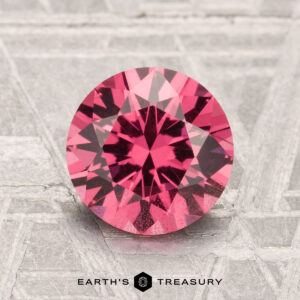 A 2.02-carat round-cut Mahenge Garnet is displayed on a textured metallic surface with "EARTH'S TREASURY" at the bottom.