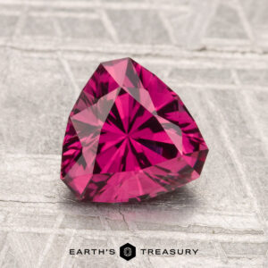A vivid, trillion-cut 1.62-carat Mahenge Garnet gemstone displayed on a textured gray surface with the "Earth's Treasury" logo below.