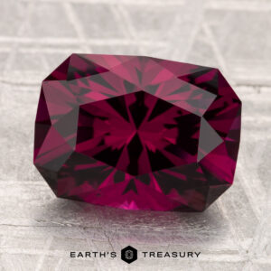 A 4.90-carat Umbalite garnet, featuring a faceted deep red hue, is showcased on a textured light gray surface with "Earth's Treasury" at the bottom.