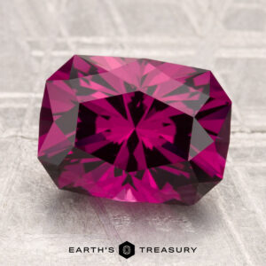 A faceted, rectangular 4.26-carat Umbalite Garnet with a rich magenta hue rests on a textured, light gray surface. The "Earth's Treasury" logo is displayed at the bottom.