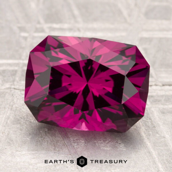 4.26-Carat Umbalite Garnet A faceted, rectangular 4.26-carat Umbalite Garnet with a rich magenta hue rests on a textured, light gray surface. The "Earth's Treasury" logo is displayed at the bottom.