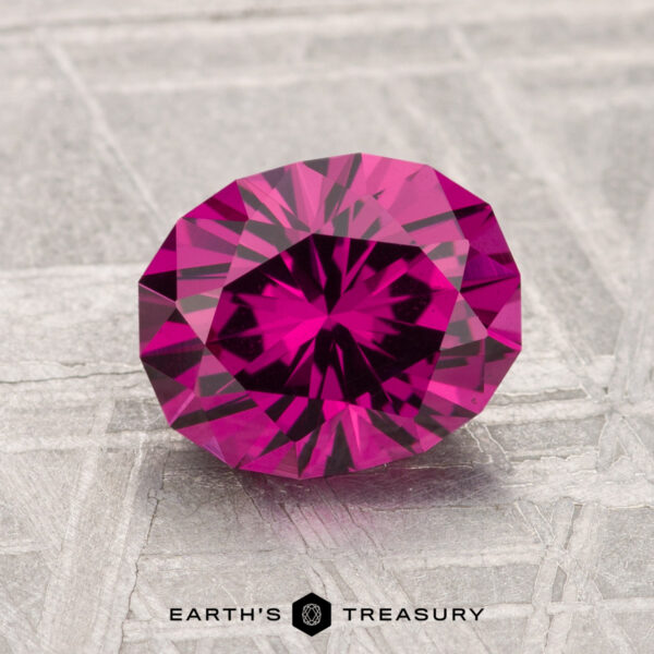 1.98-Carat Umbalite Garnet A 1.98-carat Umbalite Garnet, faceted in deep pink, rests on a textured gray surface with the text "EARTH'S TREASURY" below.