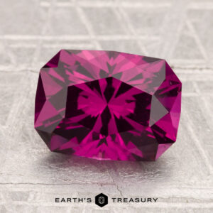 A 3.43-carat Umbalite Garnet, rectangular and faceted, rests on a textured gray surface with "EARTH'S TREASURY" visible below the gem.