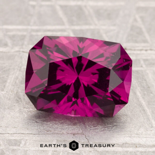 3.43-Carat Umbalite Garnet A 3.43-carat Umbalite Garnet, rectangular and faceted, rests on a textured gray surface with "EARTH'S TREASURY" visible below the gem.