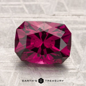 A 3.10-carat Umbalite Garnet in deep pink rests on a textured gray surface, with "Earth's Treasury" and a hexagon logo at the bottom.